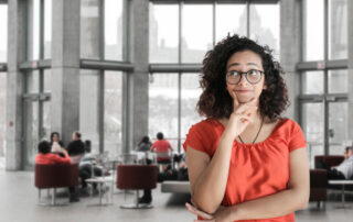 Making Decisions with the Six Hat Method image shows a woman with glasses and curly hair in the right foreground with one hand on her chin, thinking. The background shows a blurred room with chairs and people sitting or talking in front of tall windows.