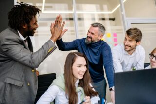 5 Reasons You Should Hire a Business Coach. Two men high five next to a smiling woman on a computer. A fourth man watches, smiling.