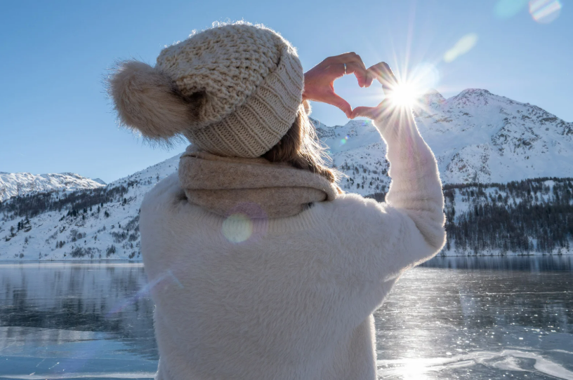 Build the Life You Want with Small Goals - image shows a woman in a sweater, scarf, and sweater facing away from the camera toward a snow-covered mountain. She holds her hands up to form a heart with her fingers.