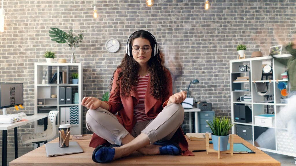 Stress Management: image shows a woman sitting on top of a desk cross-legged, headphones on, eyes closed, meditating.