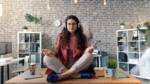 Stress Management: image shows a woman sitting on top of a desk cross-legged, headphones on, eyes closed, meditating.